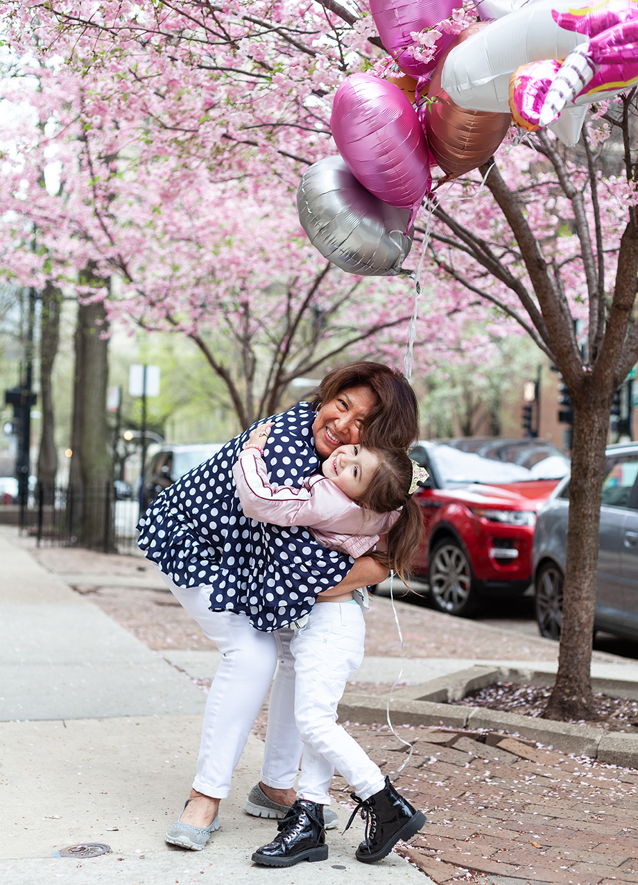 Zelda of Glitter and Bubbles hugs her nanny outside n the sidewalk while holding balloons. 