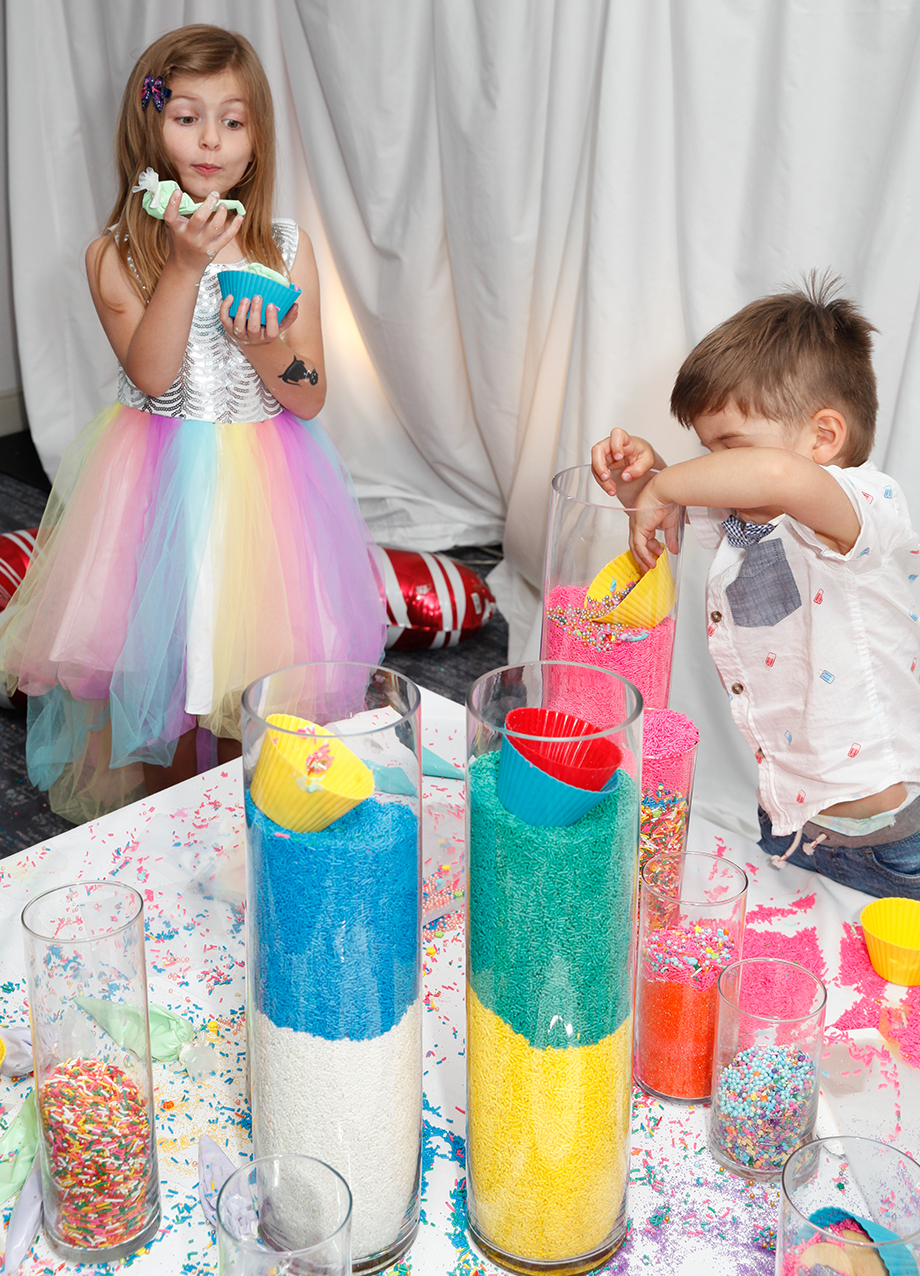 Two children decorate cookies with a giant sprinkle tower.